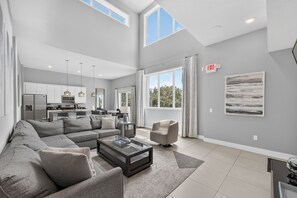 A spacious living room with a grey sectional sofa, a coffee table, and a dining area with a kitchen in the background. - 5BR Reunion Resort Condo With Elevator and Share Pools (Kissimmee)