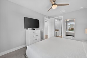 Een slaapkamer met een bed, een tv bevestigd aan een dressoir, een plafondventilator en een badkamer zichtbaar door een geopende deur.