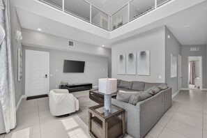 A modern living room with a high ceiling, a flat-screen TV mounted on the wall, a grey sofa, a coffee table, and a white chair. - 5BR Reunion Resort Condo With Elevator and Share Pools (Kissimmee)