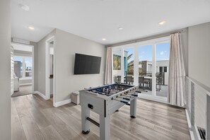 A modern room with a foosball table, a flat-screen TV, and a sliding glass door leading to an outdoor area. - 5BR Reunion Resort Condo With Elevator and Share Pools (Kissimmee)