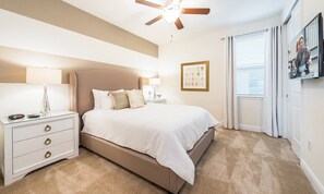 Quarto com cama, mesas de cabeceira, ventilador de teto, janela com cortinas e um quadro na parede.