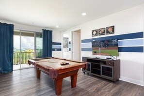 Ein Billardtisch in einem Raum mit einem Fernseher, der ein Baseballspiel zeigt, einem Balkon mit blauen Vorhängen und einer Wand mit Sportdekorationen.