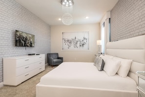A bedroom with a white bed, a grey armchair, a white dresser, and a cityscape painting on the wall. - Dainty Home With Themed Bedrooms at Encore (Kissimmee)