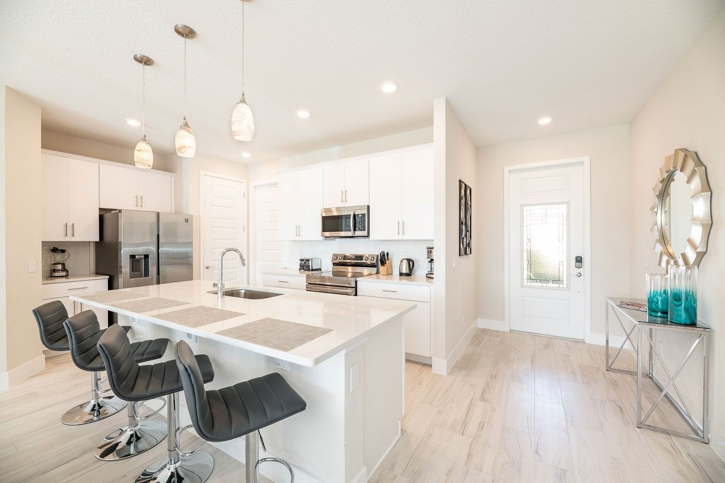 A modern kitchen with a central island, bar stools, stainless steel appliances, pendant lighting, and a decorative mirror.