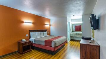 A hotel room with a wooden floor, a bed with a headboard, a bedside table with a phone, a small wooden cabinet, and a television mounted on the wall.