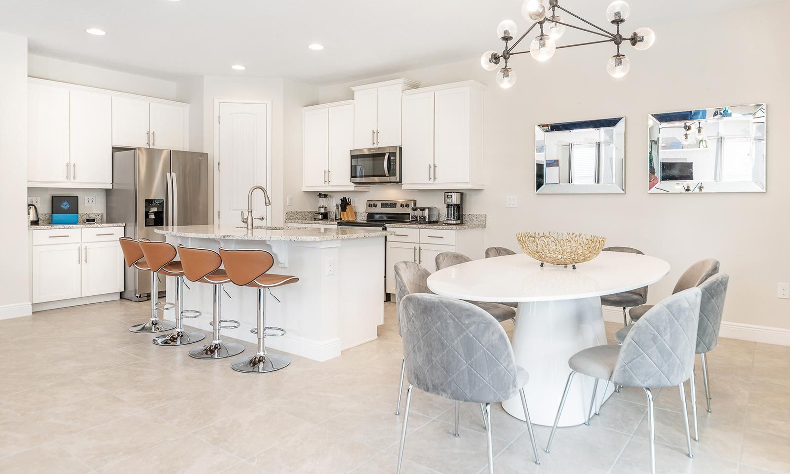 A modern kitchen with white cabinets, a stainless steel refrigerator, a microwave, and a coffee maker. There is a round dining table with grey chairs and a decorative bowl on it.