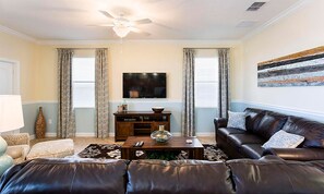 A living room with a leather couch, a coffee table, a TV, and a ceiling fan. - 6BR Championsgate Villa Pool, Spa & Game Room (Davenport)