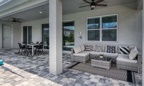 Ein überdachter Patio-Bereich mit Esstisch und Stühlen, einer Couch mit Kissen und einem Deckenventilator.