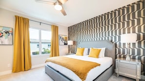 Une chambre moderne avec un grand lit, un ventilateur de plafond, une table de chevet avec une lampe et un mur décoré d’un motif.
