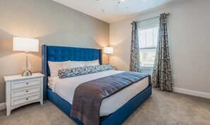 A bedroom with a blue upholstered headboard, a white bedside table with drawers, a lamp, and a window with floral curtains. - Encore 8BR Villa, Putting Green, Near Disney (Kissimmee)
