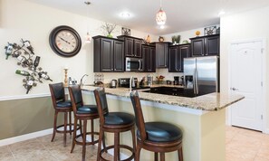 A kitchen with dark cabinets, a stainless steel refrigerator, a granite countertop, and a bar area with stools. - 7BR Championsgate Pool, Spa & Loft Games (Davenport)
