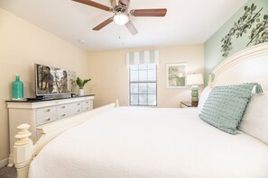 Une chambre à coucher comprenant un lit, un ventilateur de plafond, une commode, un téléviseur et une fenêtre avec des stores.