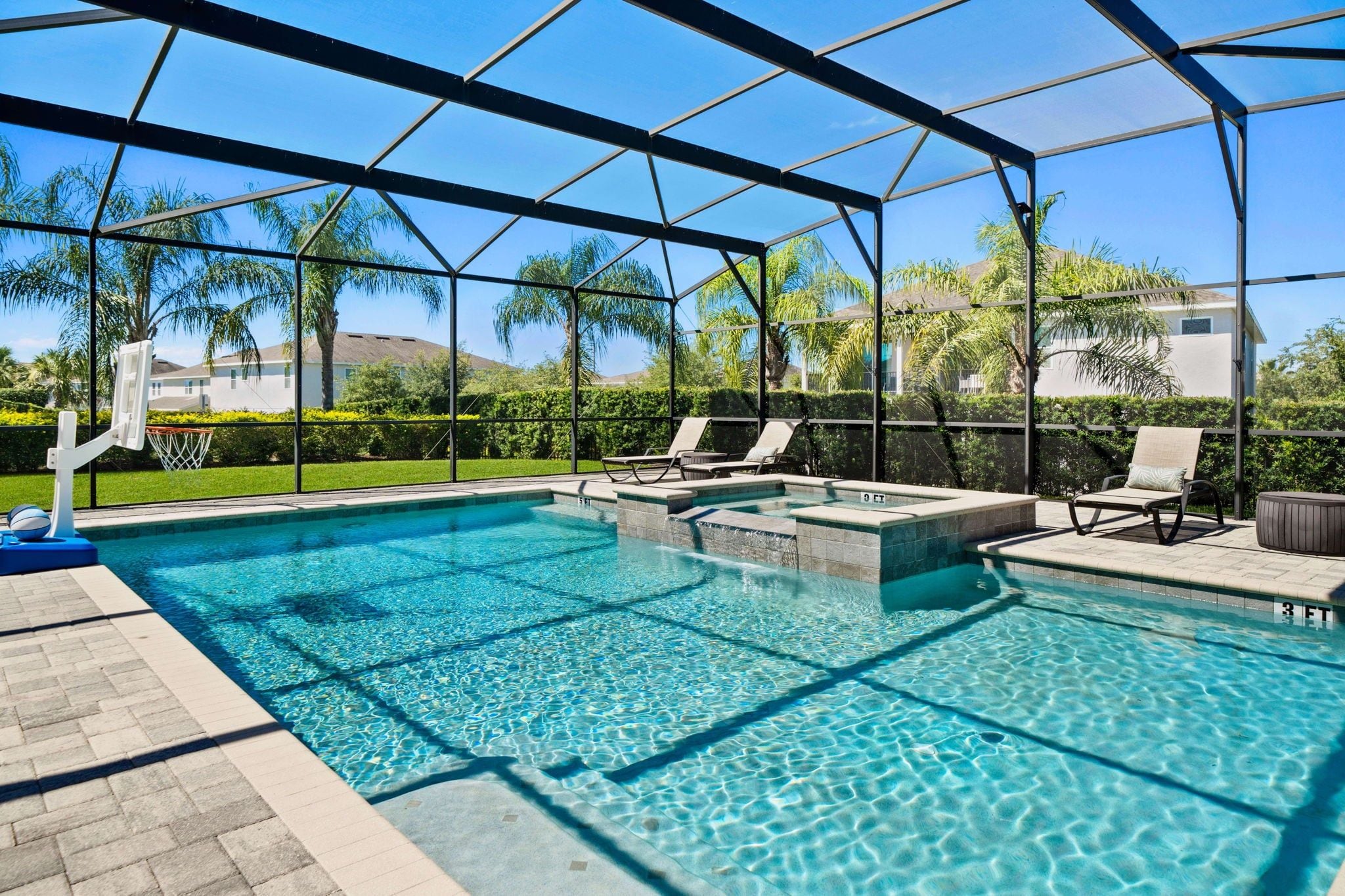 A clear swimming pool with a sunshade structure, lounge chairs, and a basketball hoop.