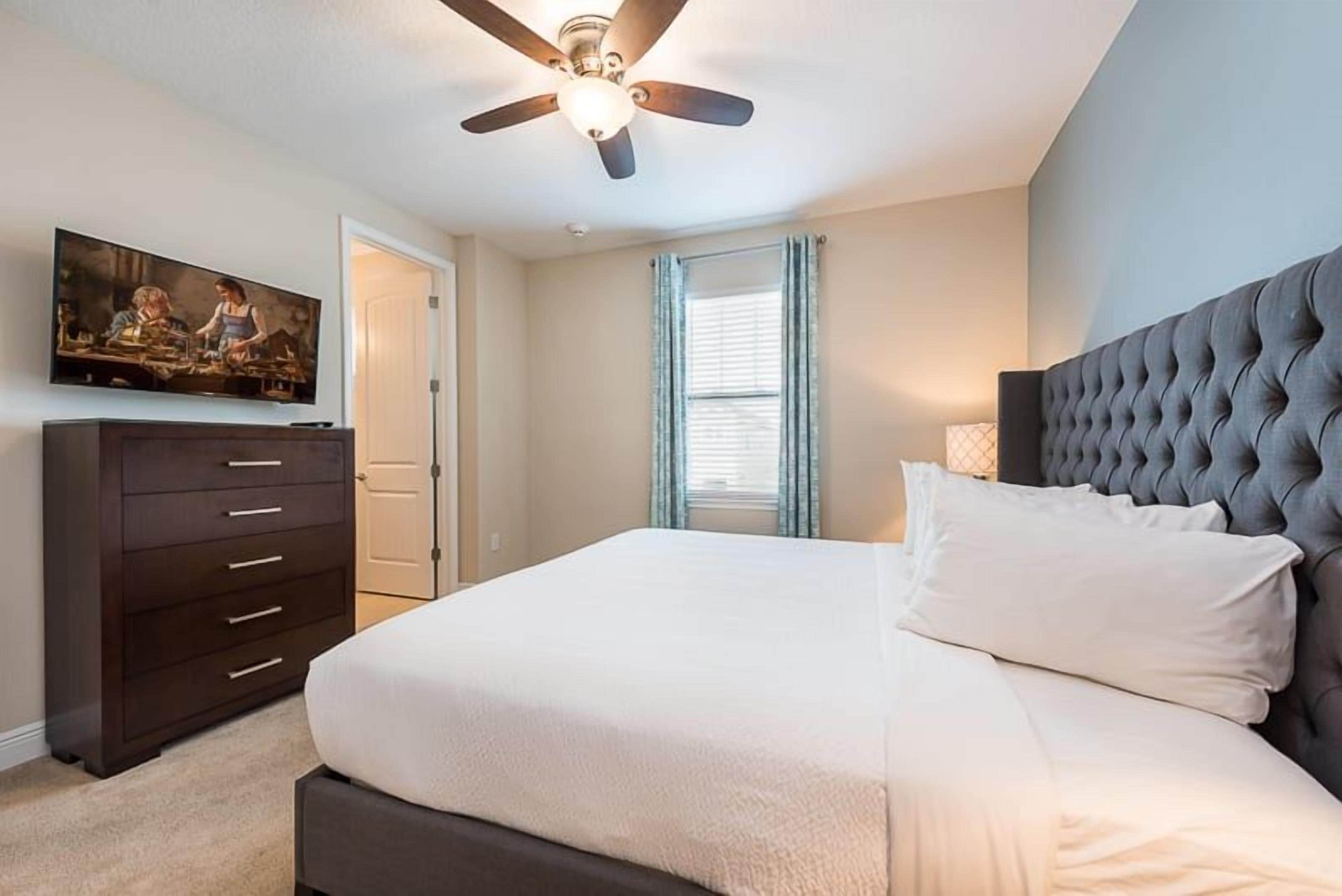 A bedroom with a bed, a dresser, a ceiling fan, a television on the wall, and a window with curtains.
