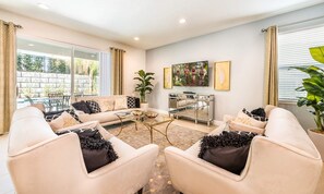 A living room with a sofa set, a coffee table, and a side table. There is a window with curtains, a potted plant, and a painting on the wall. - Delightful Home With Private Pool Oasis in Encore (Kissimmee)