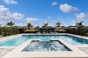 Una piscina rectangular con un jacuzzi en el centro, rodeada de sillas de descanso y palmeras.