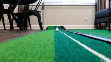 A room with a green artificial turf playing field, a white golf ball, and a black pool cue.