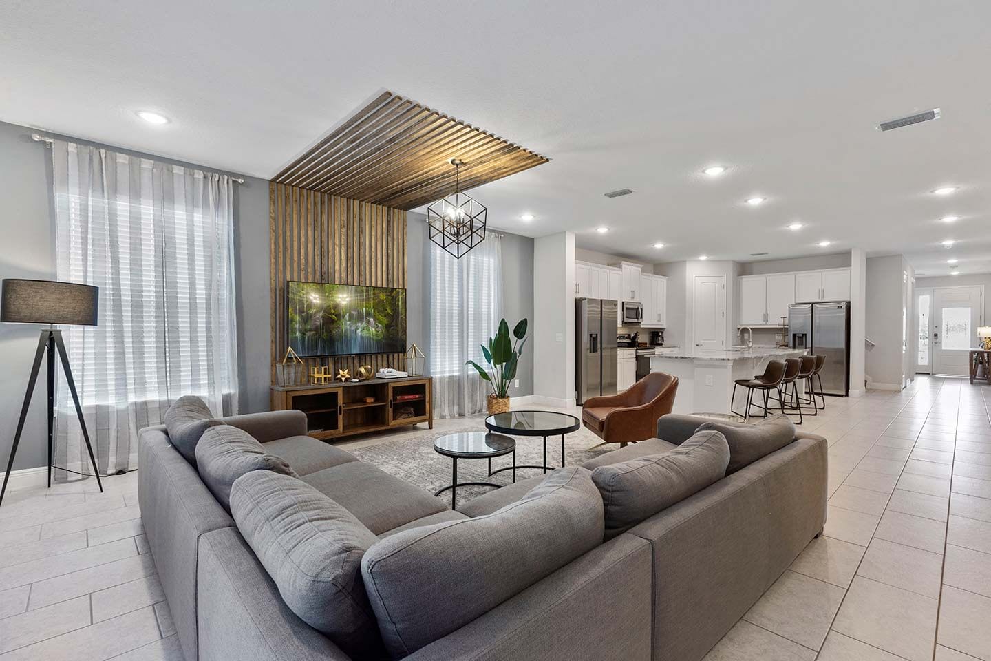 Ein modernes Wohnzimmer mit einem großen grauen Sofa, einem Fernseher auf einem Holzständer, einem kleinen runden Tisch und einer Küche im Hintergrund.