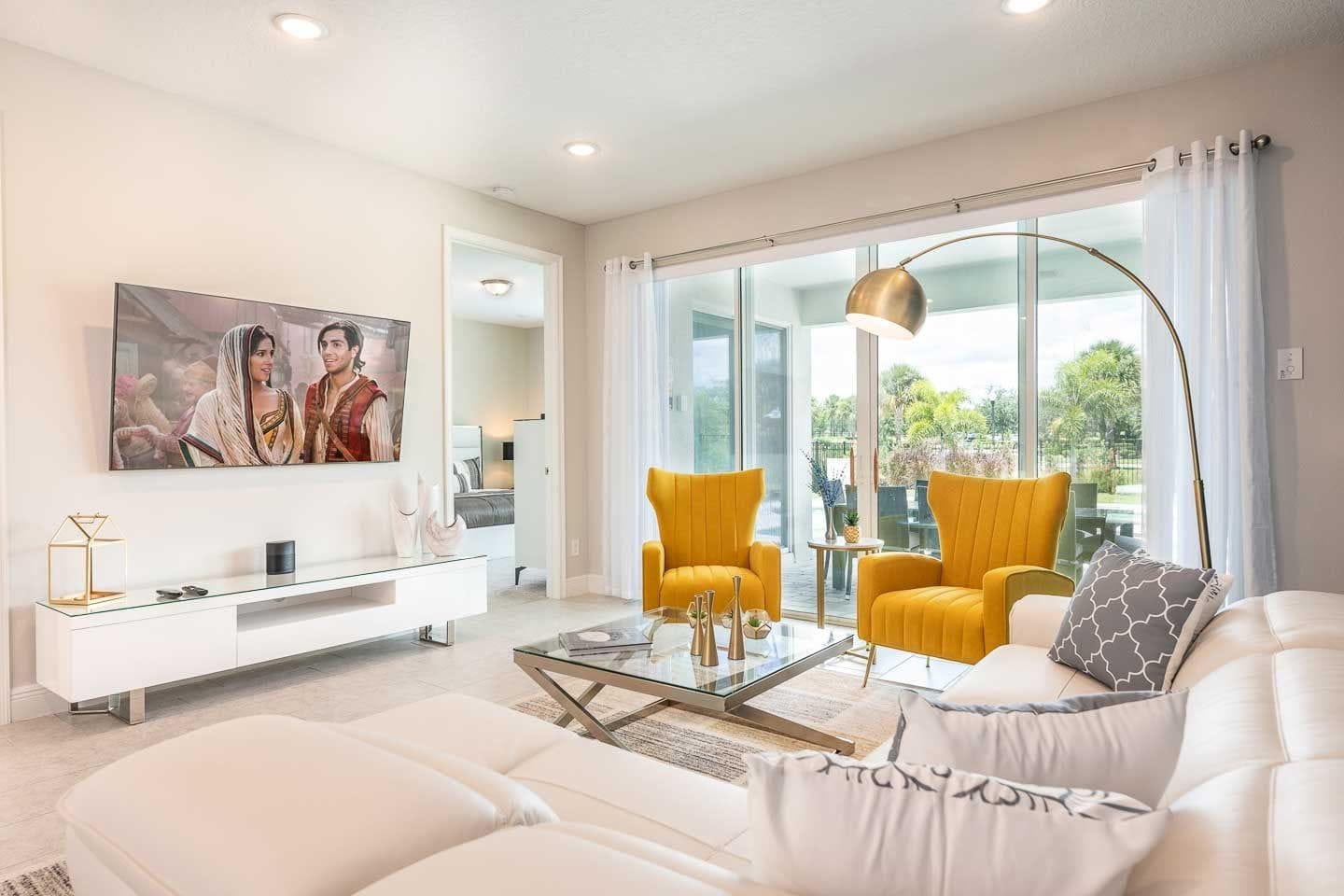 A modern living room with a white sofa, yellow armchairs, a glass coffee table, and a flat-screen TV mounted on the wall.