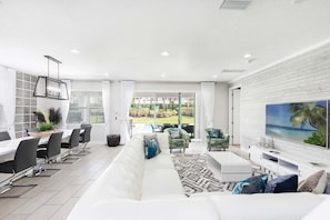 Ein modernes Wohnzimmer mit einem weißen Sofa, einem Essbereich mit einem langen Tisch und Stühlen, einem an einer weißen Ziegelwand montierten Fernseher und Blick auf einen Garten durch große Fenster.