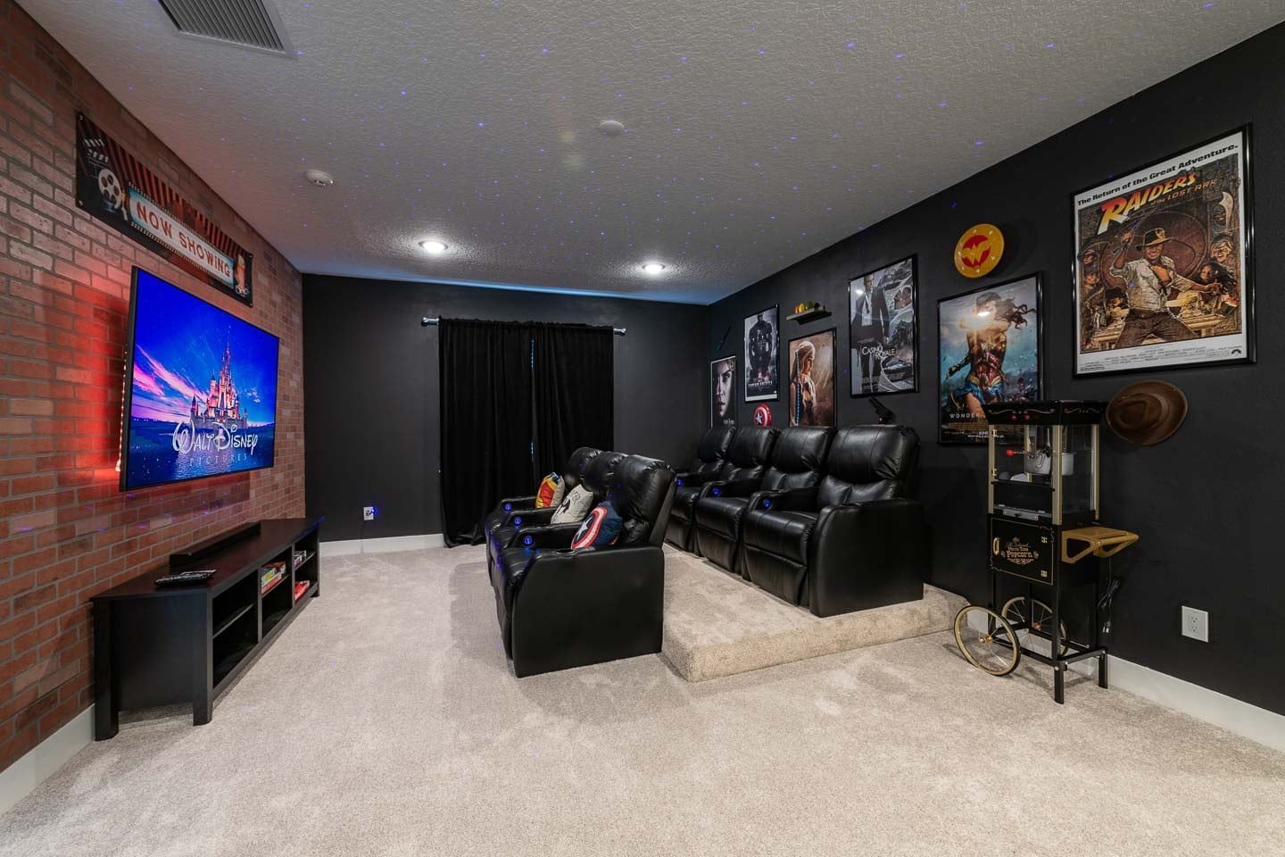 A home theater room with a flat-screen TV, black leather recliners, a popcorn machine, and movie posters on the walls.