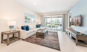 A modern living room with a sofa, coffee table, and TV. There is a balcony with a view of the outside. - Captivating Villa With Theater at Encore (Kissimmee)