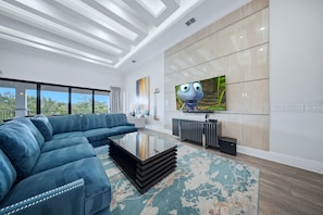 A modern living room with a sectional sofa, a coffee table, a flat-screen TV, and a large window offering a view of the outdoors. - Deluxe Mansion With Swim up Bar and Pool Slide at Reunion (Kissimmee)