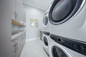 A laundry room with two stacked front-loading washing machines, a countertop with a sink, and a wall-mounted drying rack. - Deluxe Mansion With Swim up Bar and Pool Slide at Reunion (Kissimmee)
