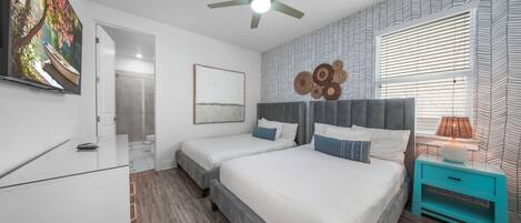 Un dormitorio con cama, mesitas de noche, ventilador de techo, ventana con persianas y un cuadro en la pared.