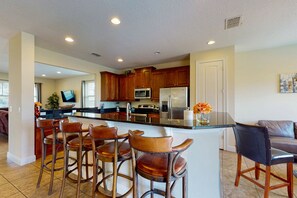 A modern kitchen with wooden cabinets, a stainless steel refrigerator, and a bar area with leather stools. - Charming 6BR Encore Villa Pool, Spa & Disney Near (Kissimmee)