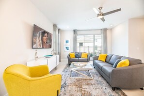 A modern living room with a sofa, armchair, and coffee table, a ceiling fan, and a large window with curtains. - Bright and Sunny Townhouse With Games Room at Reunion (Kissimmee)