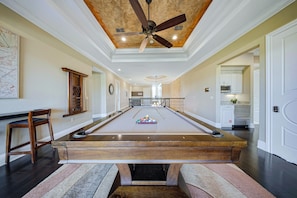 A spacious room with a pool table, a ceiling fan, and a kitchen area visible through an open doorway. - Delightful Haven With Theater Room at Reunion (Kissimmee)