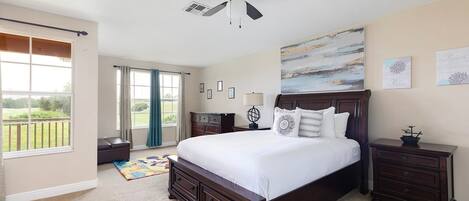 A bedroom with a large bed, a ceiling fan, a view through a window, and a painting on the wall.