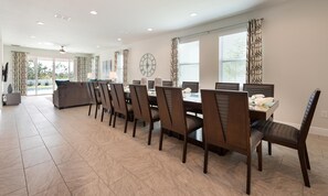 A spacious dining area with a long table and multiple chairs, a clock on the wall, and large windows with curtains. - Spacious Abode With Spillover Spa in Encore (Kissimmee)