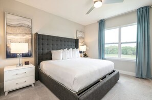 A modern bedroom with a large bed, a nightstand, a lamp, and a view of greenery through the window. - The Tranquil Townhome - Elegant Townhome in Reunion Resort (Kissimmee)