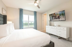 A bedroom with a large bed, a ceiling fan, a white dresser, a painting, and a window with curtains. - The Tranquil Townhome - Elegant Townhome in Reunion Resort (Kissimmee)