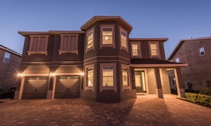 A two-story house with a garage, a covered entryway, and a well-lit driveway at dusk. - Luxurious Villa With Tiki Themed Game Room at Encore (Kissimmee)