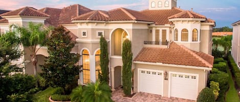 A large, multi-story house with a tiled roof, multiple garage doors, and a well-maintained garden with various plants and a paved driveway.