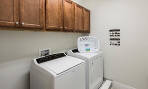 A laundry area with a washing machine and dryer, wooden cabinets above, and laundry instructions on the wall. - Sun-kissed Villa With Private Pool at Encore (Kissimmee)