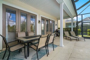 A covered patio with a table and chairs, large windows with blinds, and a grill. - Tropical Retreat With Screened in Private Pool and Spa at Reunion (Kissimmee)