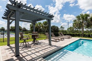 A poolside area with a pergola, lounge chairs, and a table. - Energetic Villa With Gazebo at Encore (Kissimmee)