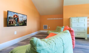 Ein Zimmer mit einem großen Flachbildfernseher an der Wand, einem weißen Schminktisch und zwei grünen Sitzsäcken mit orangenen Kissen.