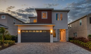 A modern two-story house with a garage, stone accents, and a well-lit entrance. - Modern 4 Bedroom Home With Game Room in Encore Resort (Kissimmee)