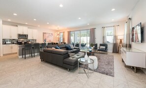 A modern living room with a sectional sofa, a dining area with a kitchen in the background, and large windows providing natural light. - Modern 4 Bedroom Home With Game Room in Encore Resort (Kissimmee)
