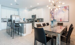 A modern kitchen with white cabinets, a central island, and a dining area with a glass table and black chairs. - Modern 4 Bedroom Home With Game Room in Encore Resort (Kissimmee)