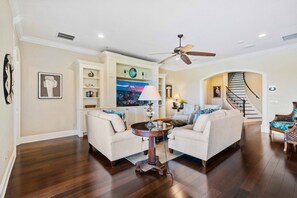 A spacious living room with a high ceiling, a ceiling fan, a large window with a view, and a staircase leading to an upper level. - Noble Estate With Private Pool at Reunion (Kissimmee)