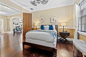A spacious bedroom with a large bed, two bedside lamps, a wooden headboard, and a view of a hallway with a door and a chair. - Noble Estate With Private Pool at Reunion (Kissimmee)