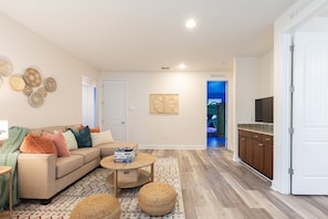 A living room with a sofa, coffee table, and ottomans, a kitchen with cabinets and a TV, and a door leading to a bedroom. - Relax at Encore: 8br, Pool, Minutes to Disney (Kissimmee)