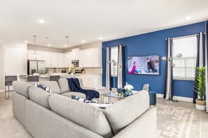 A modern living room with a grey sofa, a flat-screen TV, and a kitchen in the background. - Sophisticated Villa With Movie Room at Encore (Kissimmee)