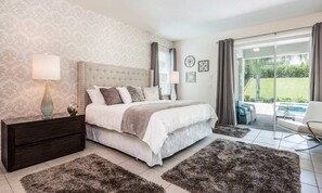 A bedroom with a large bed, a nightstand, a lamp, and a view of a garden through a sliding glass door. - Stylish Sleek Home With Full Kitchen at Encore (Kissimmee)